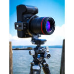 Leofoto UL-03 Objectiefvoet in gebruik met camera en telelens in verticale stand op statief bij een meer onder een heldere lucht.