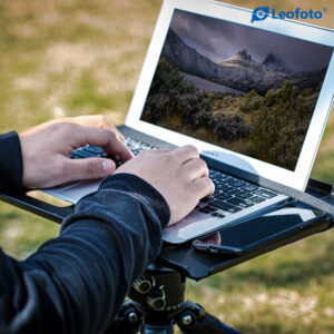 Leofoto LCH-2S laptophouder gebruiksvoorbeeld buiten met laptop op tray op statief terwijl de gebruiker typt.